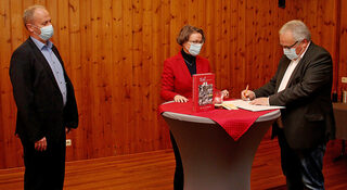 Uwe Schubinski (r.) unterzeichnet unter den Augen von Ministerin Scharrenbach und Bürgermeister Esser den Förderbescheid.